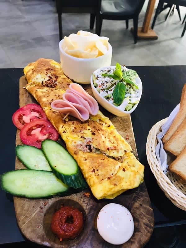 Long seasoned omelet on a wooden board with ham, cucumbers, tomatoes, salad, and toast.